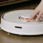 Close-up of a hand pressing a button on a robotic vacuum cleaner on a wooden floor.
