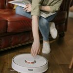 Person relaxing in cozy living room while operating a robot vacuum cleaner on wooden floor.